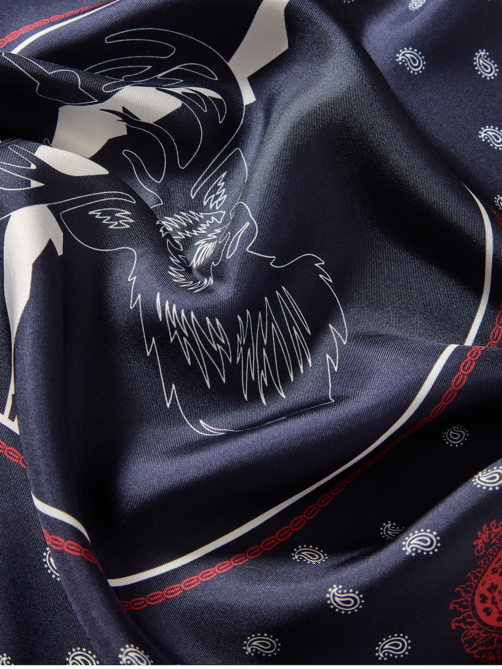 A close-up of the We Norwegians Western Silk Scarf in dark blue, featuring a white stag’s head with antlers and red-and-white paisley bandana borders—softly folded to reflect Norwegian heritage.