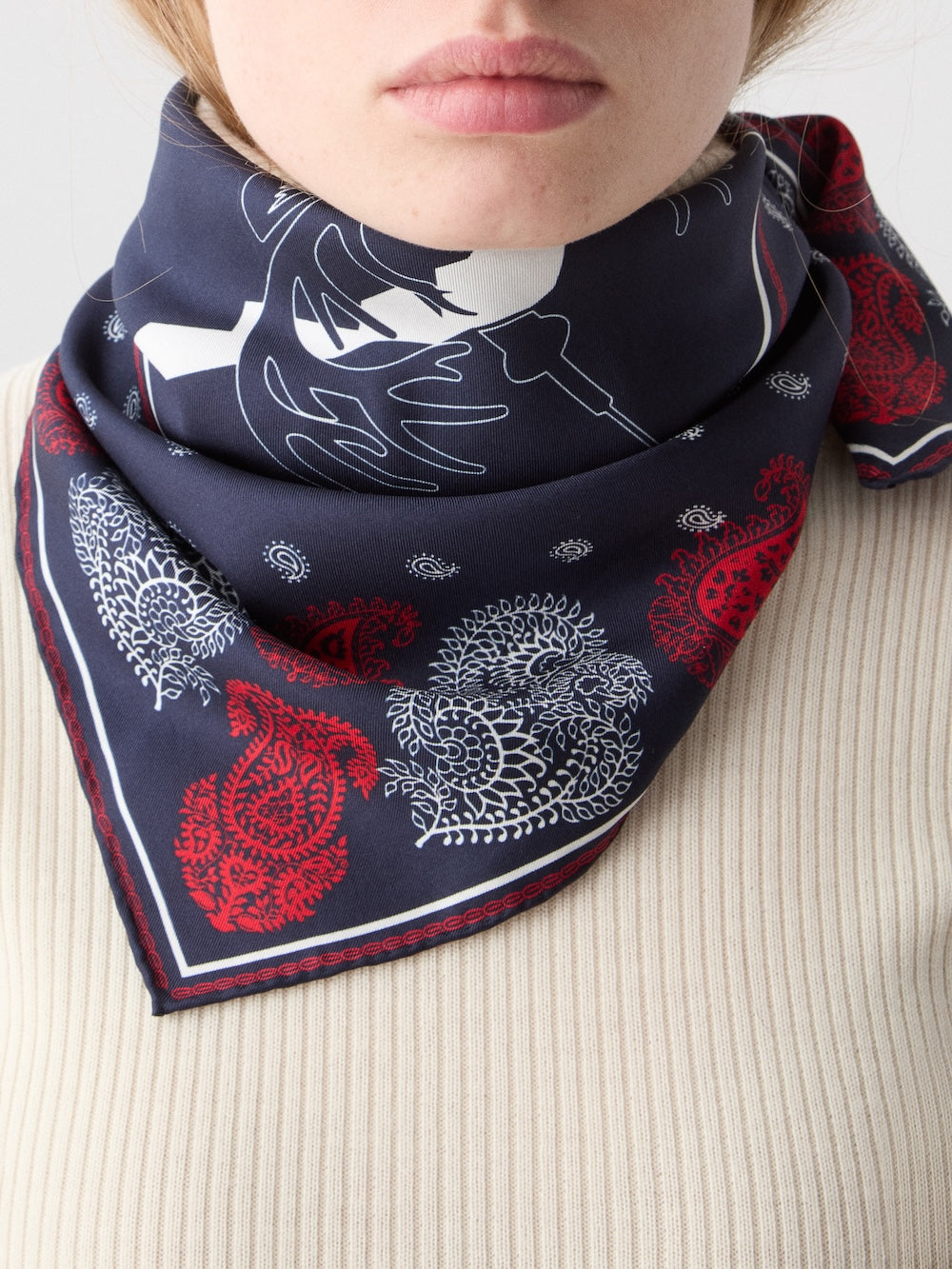 A person wears the Western Silk Scarf by We Norwegians, featuring white and red paisley patterns inspired by Norwegian heritage, paired with a ribbed beige top. Only the lower half of the face and neck are visible.