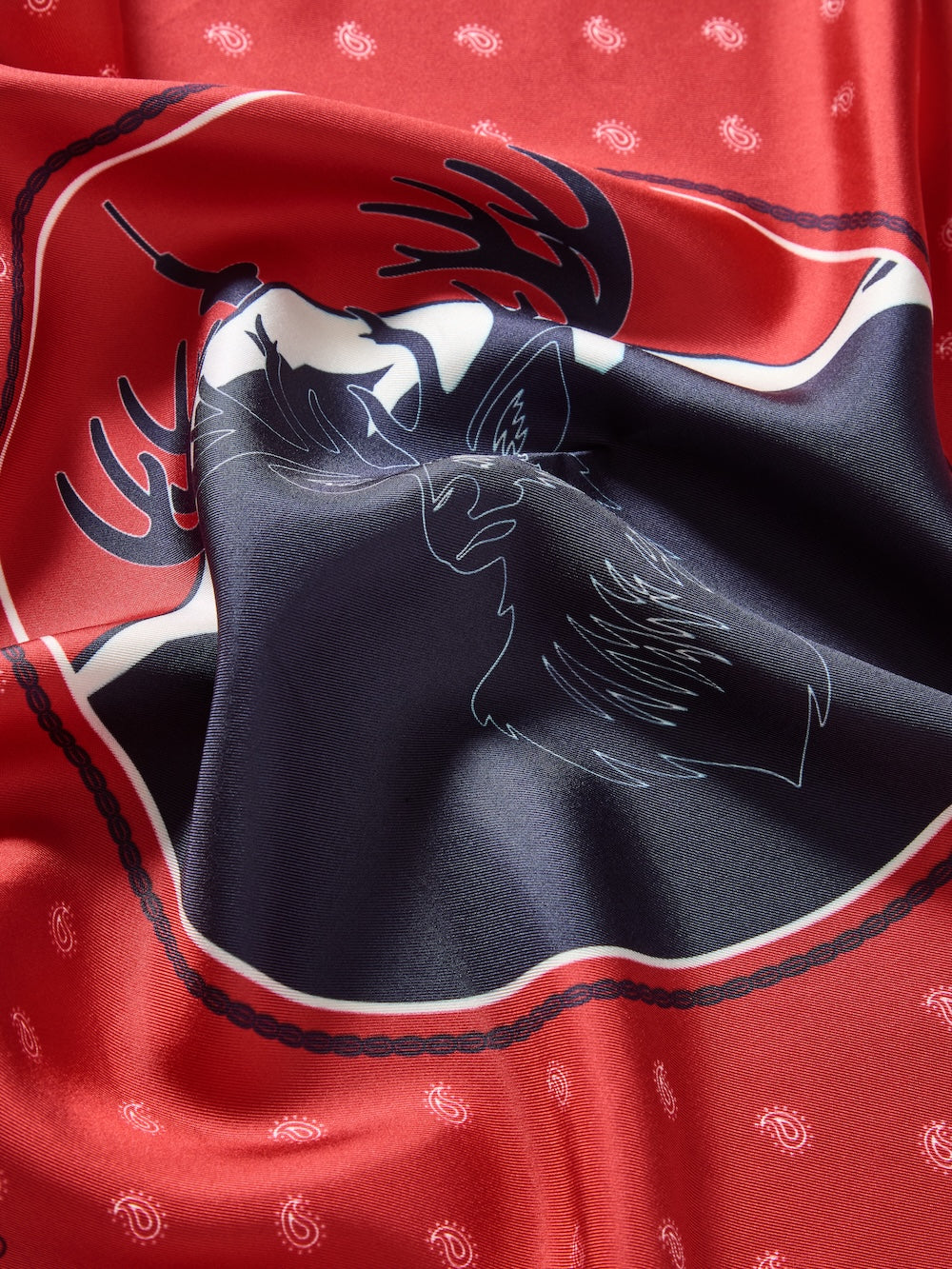 A close-up of the We Norwegians Western Silk Scarf in shiny red, styled like a bandana, shows a black deer silhouette with antlers outlined in white and a paisley pattern along the softly crumpled edges.