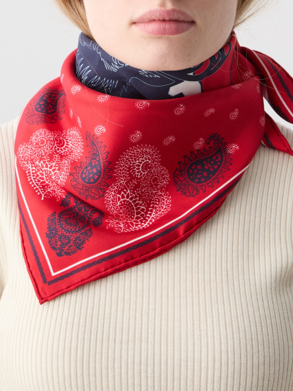 A person wearing a cream ribbed top and the We Norwegians Western Silk Scarf, a red paisley bandana with white and navy details, tied at the neck. Only their lower face and neck are visible, evoking classic Norwegian heritage.