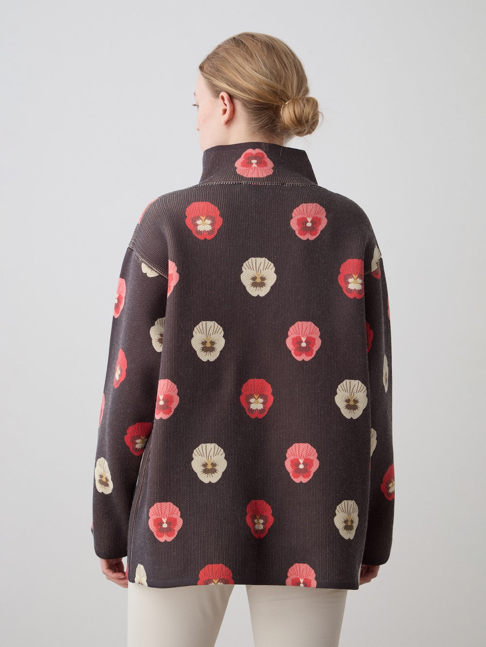 A woman with light brown hair in a low bun faces away, wearing the We Norwegians WN x Markarian Jacket for women—a dark, high-collared piece with a red and beige floral pattern—against a plain, light gray background.