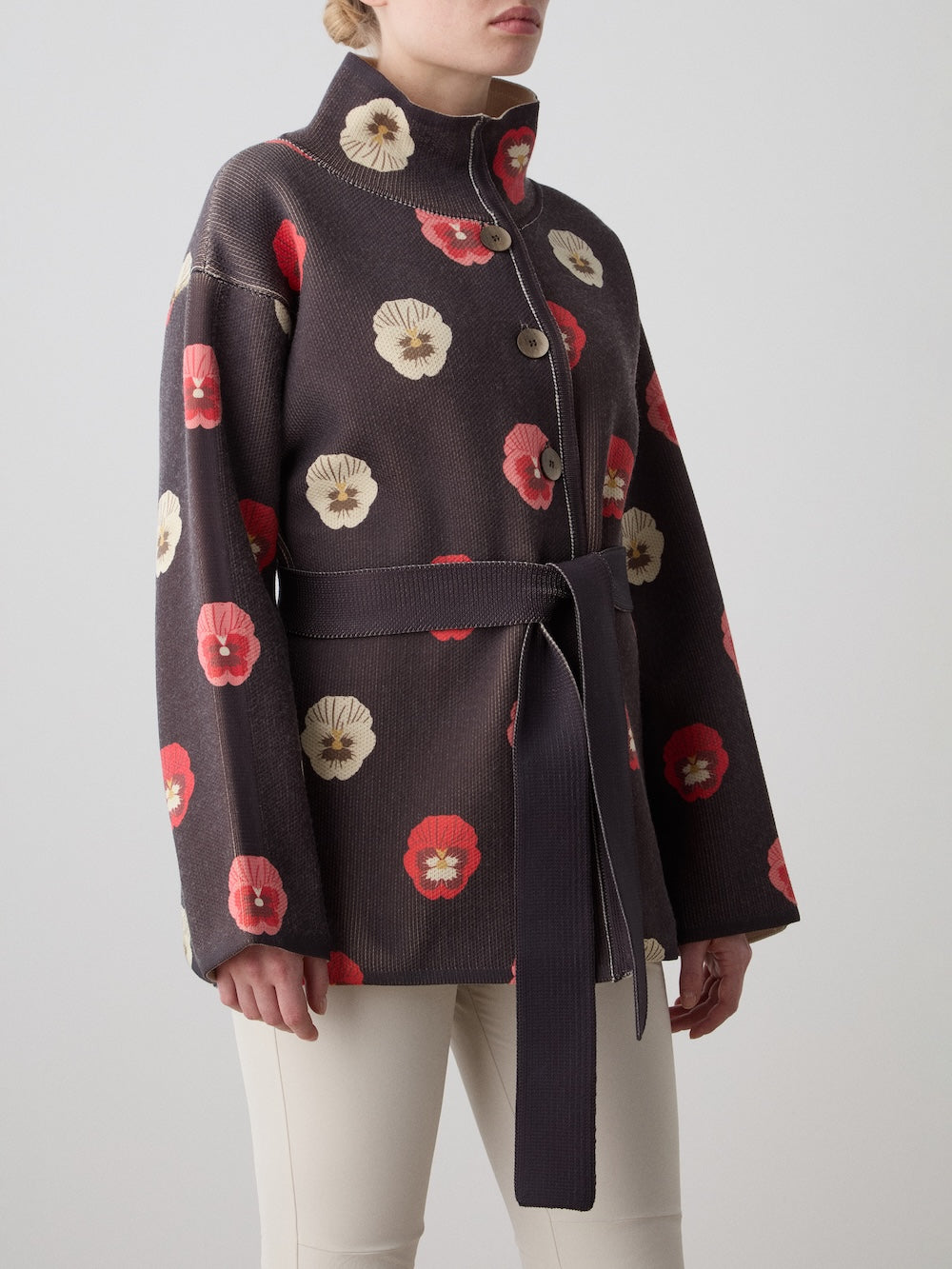A person is seen in a WN x Markarian Jacket by We Norwegians, featuring a red and white floral print with matching belt, styled with light pants; only their lower face and body are shown.