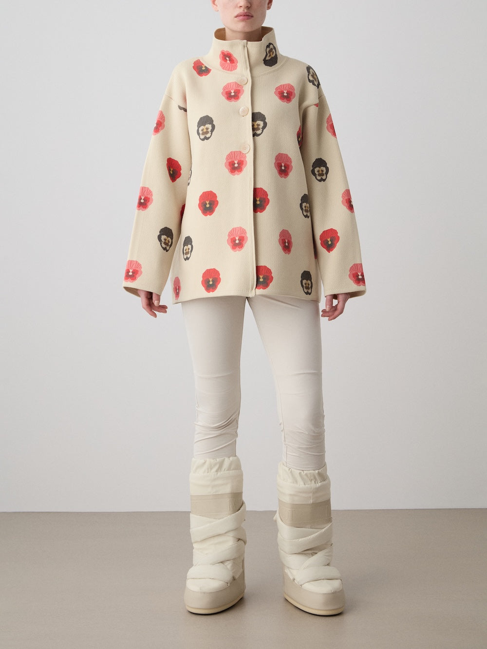 A person models the We Norwegians WN x Markarian Jacket for women, featuring digitally printed wool with red and black floral patterns, styled with fitted cream pants and chunky quilted cream boots against a plain backdrop.