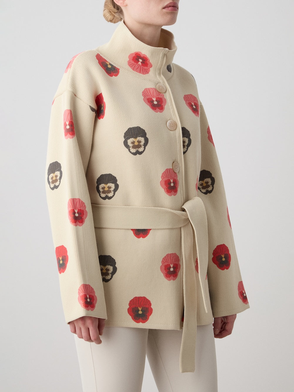 A woman models the limited edition WN x Markarian Jacket by We Norwegians, featuring red and dark floral prints on beige digitally printed wool with a belt, large buttons, and a high collar, against a plain light background.