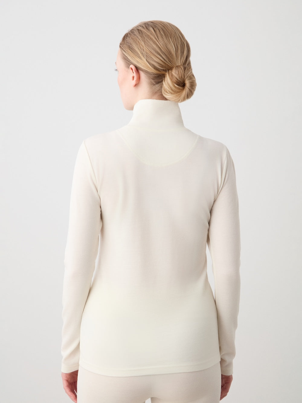 A person with blonde hair in a low bun is seen from the back, wearing the cream-colored WN x Markarian ZipUp women base layer by We Norwegians, against a plain light background.
