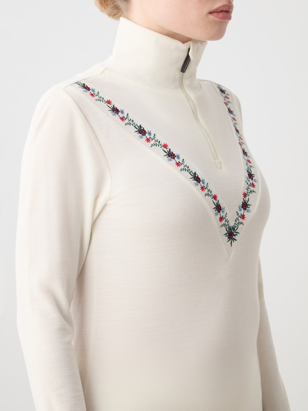 A woman wears the We Norwegians WN x Markarian ZipUp, a cream long-sleeve merino wool base layer with a high zip collar and V-shaped red, black, and green floral embroidery on the chest.