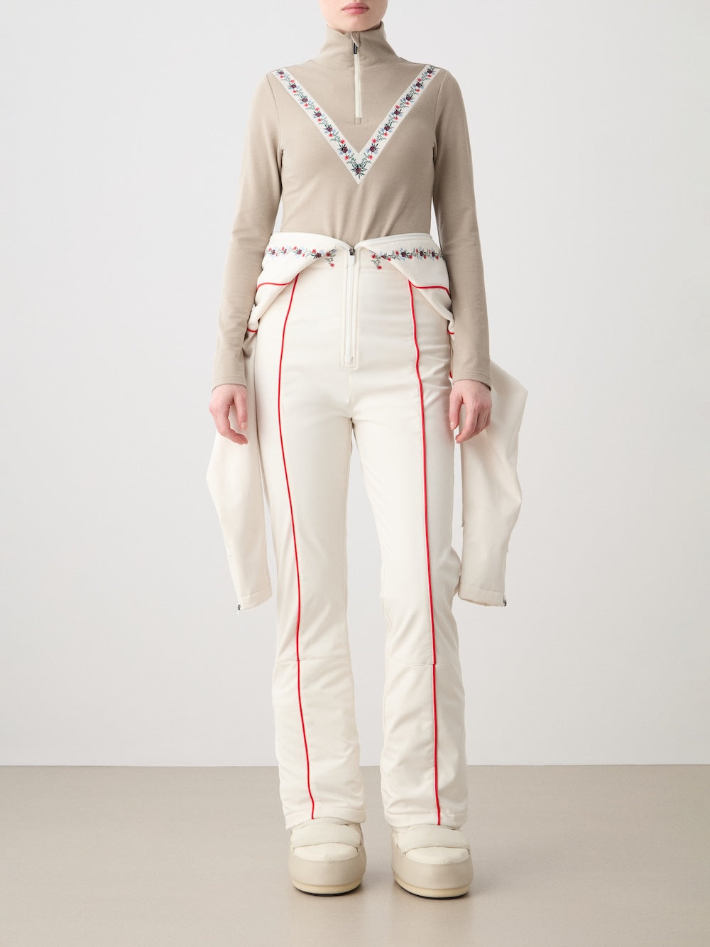 A person faces forward in a We Norwegians WN x Markarian ZipUp women’s beige top with colorful embroidery, high-waisted white pants with red piping, and cream boots, set against a plain neutral background.