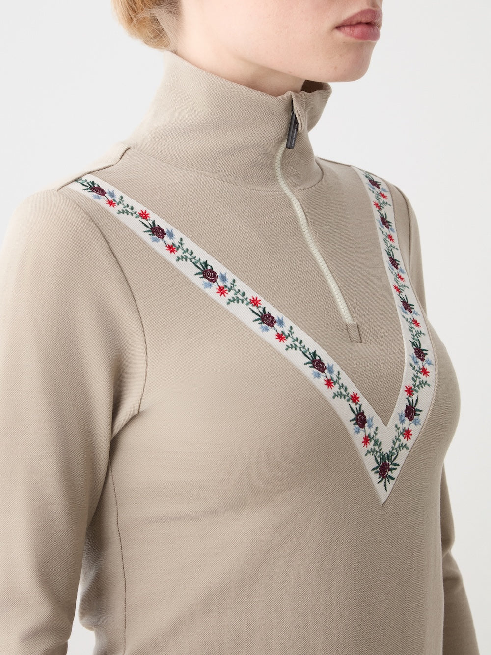 A close-up of someone wearing the We Norwegians WN x Markarian ZipUp women’s base layer, featuring a beige merino wool fabric, zippered high collar, and a V-shaped red, blue, green, and white floral embroidery across the chest and shoulders.