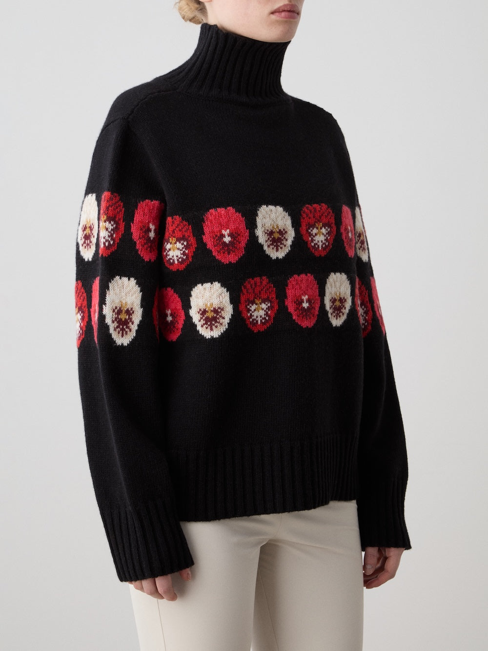 A person wears the limited edition WN x Markarian Sweater for women by We Norwegians, featuring red, white, and pink Fair Isle floral patterns on the chest and sleeves, paired with light pants against a plain background.