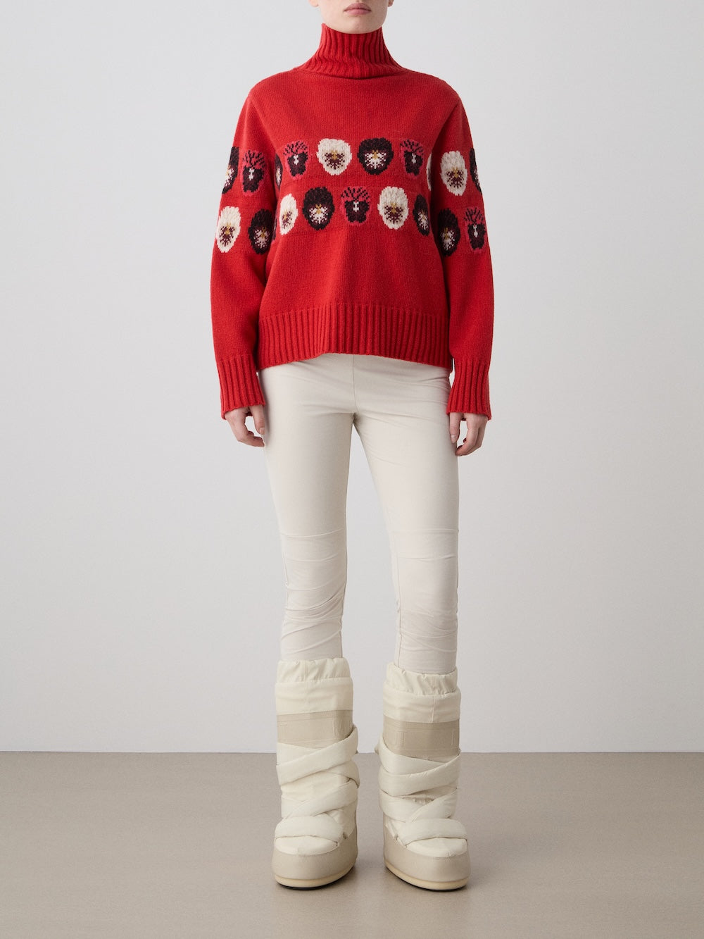 A person stands by a plain wall in a limited edition red WN x Markarian Sweater by We Norwegians, paired with off-white fitted pants and tall, puffy off-white winter boots. Their face is not shown.