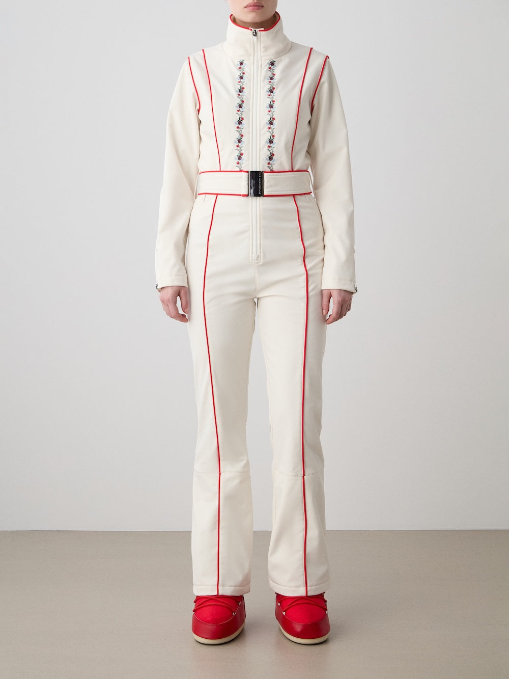 A person stands against a plain background wearing the We Norwegians WN x Markarian Ski Jumpsuit for women, featuring red piping, floral chest embroidery, a wide black belt, and red snow boots. Their hands rest relaxed at their sides.