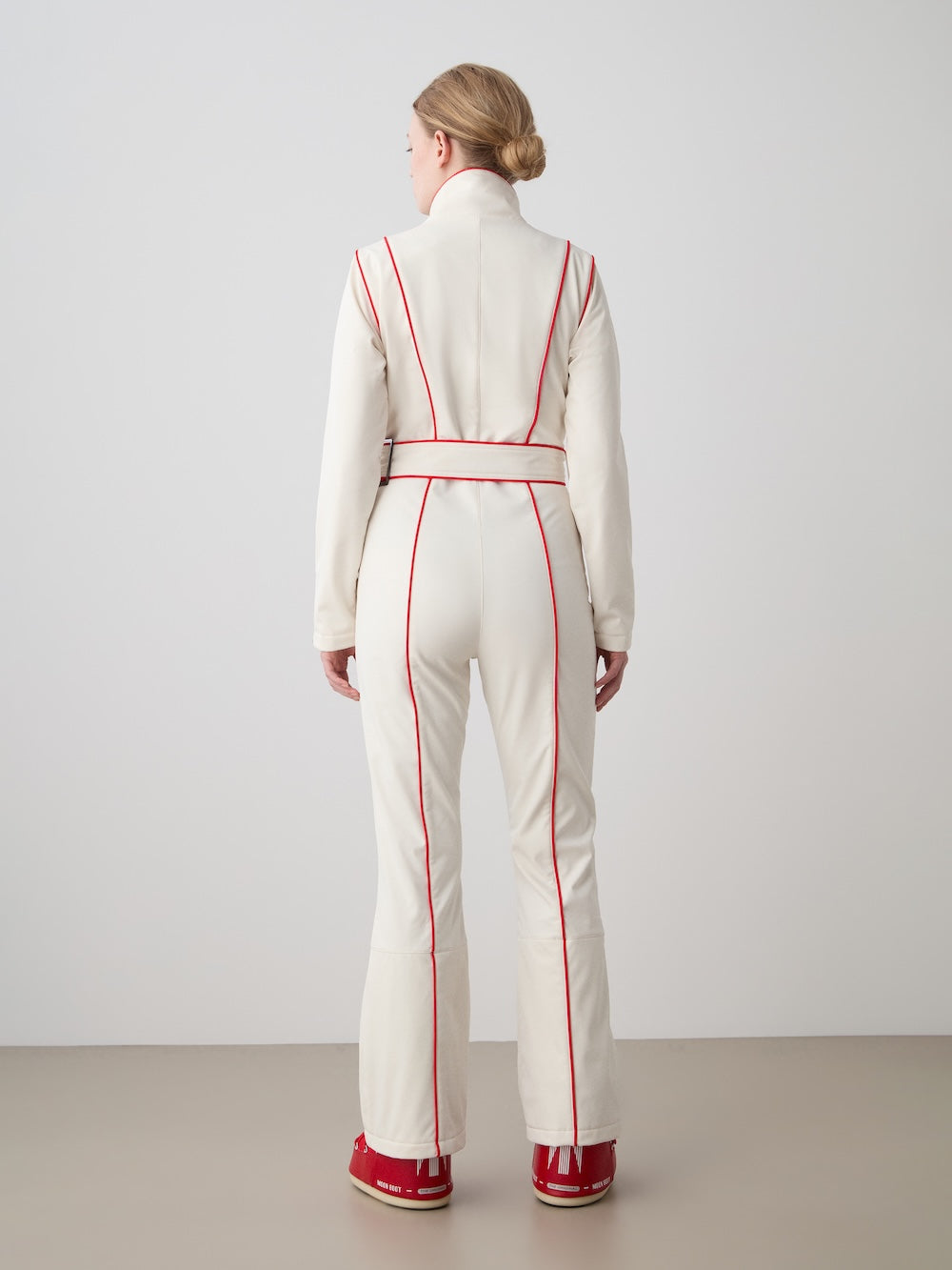 A woman with blond hair in a low bun, wearing the fitted white We Norwegians WN x Markarian Ski Jumpsuit with red vertical piping and red shoes, stands facing away against a plain light gray background.