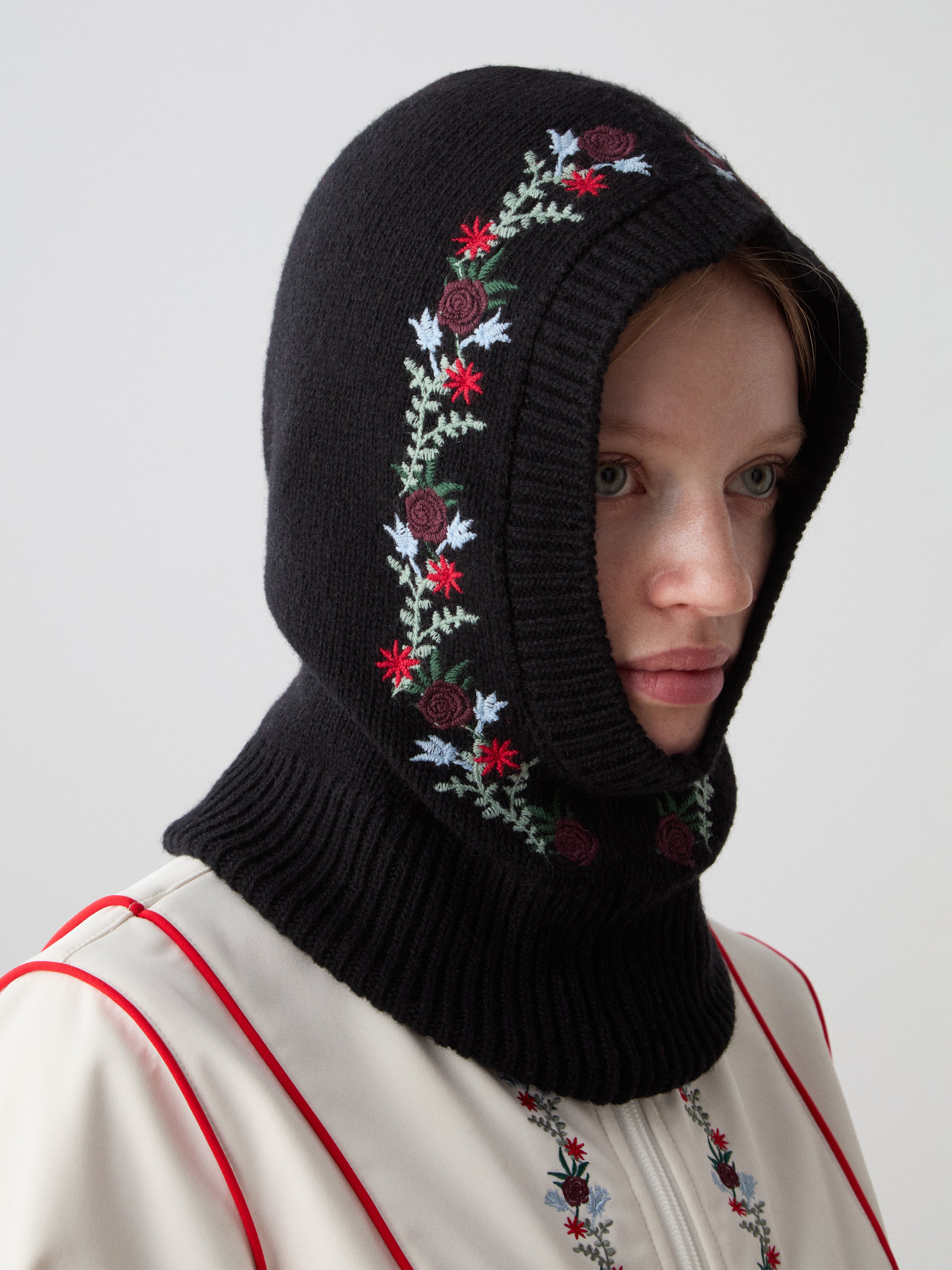 A woman models the limited edition We Norwegians WN x Markarian Balaclava, featuring colorful floral embroidery, with a cream top with red and blue stitching, gazing slightly right against a plain background.