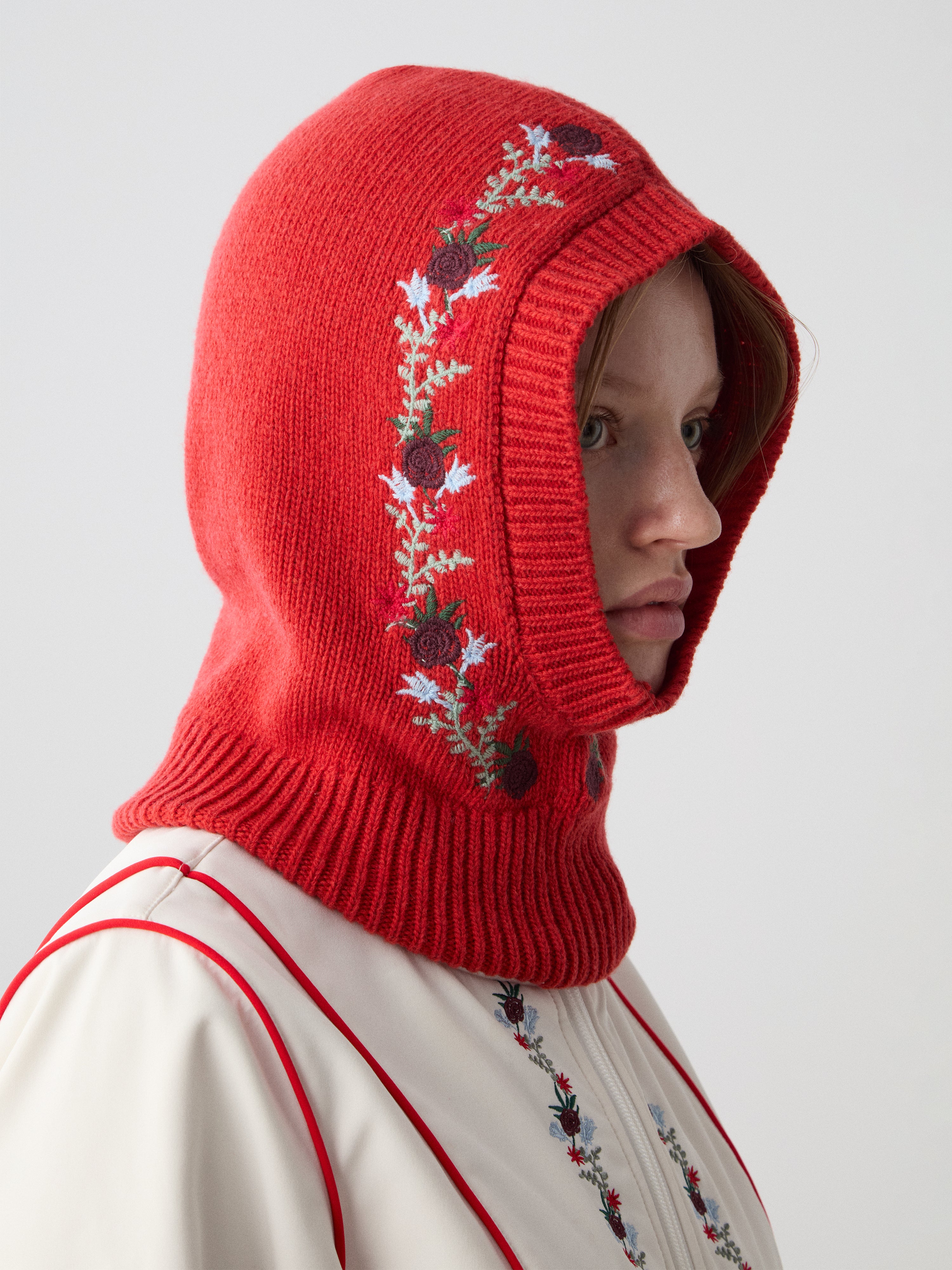 A woman wears the limited edition WN x Markarian Balaclava by We Norwegians, a red wool piece with floral embroidery, paired with a cream embroidered garment against a light background.
