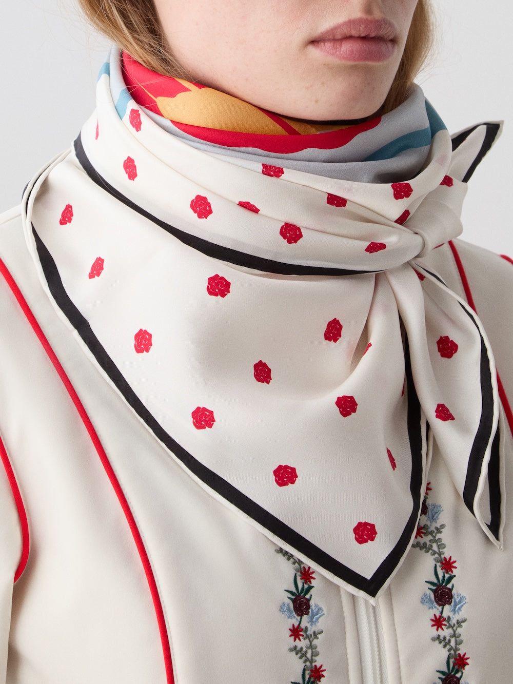 A woman wears the WN x Markarian Silk Scarf women by We Norwegians, a limited-edition white silk scarf with red floral dots and black trim, loosely tied around her neck. She pairs it with a cream top featuring red piping and floral embroidery.
