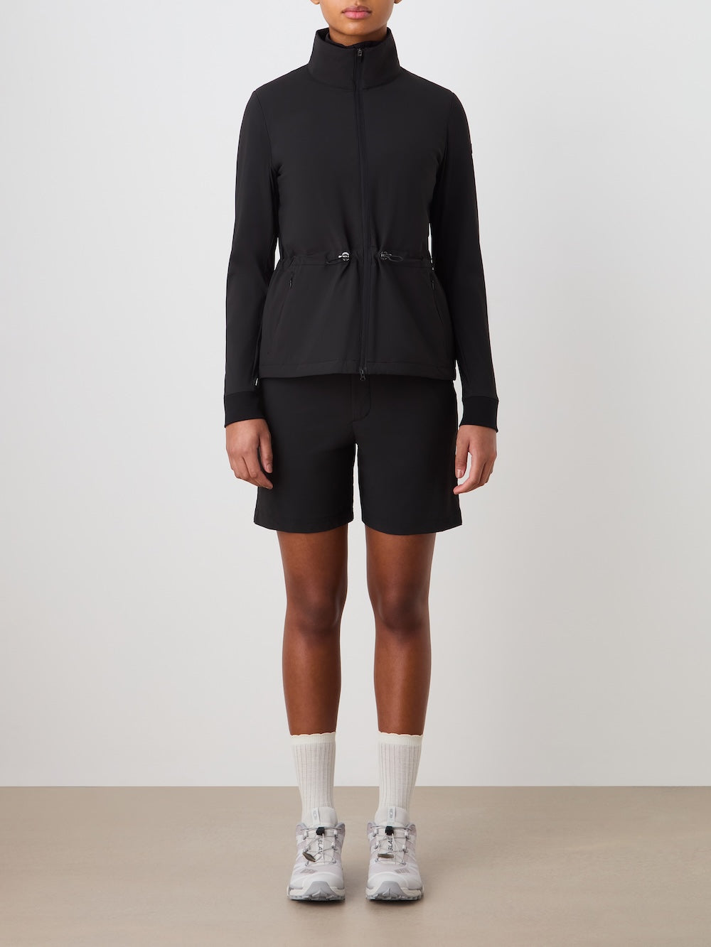 A person stands against a plain background wearing the We Norwegians Albatross Jacket Women, which is black, water-repellent, and made with stretch fabric. They pair it with black shorts, white crew socks, and white sneakers for a sporty, casual look.