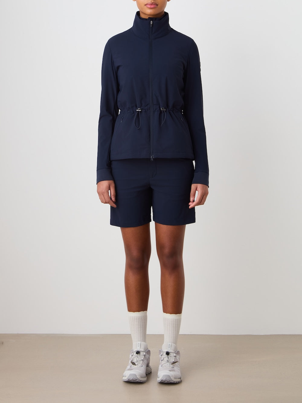A person faces forward against a plain background, wearing the We Norwegians Albatross Jacket Women in navy with matching shorts, white crew socks, and light gray athletic shoes.