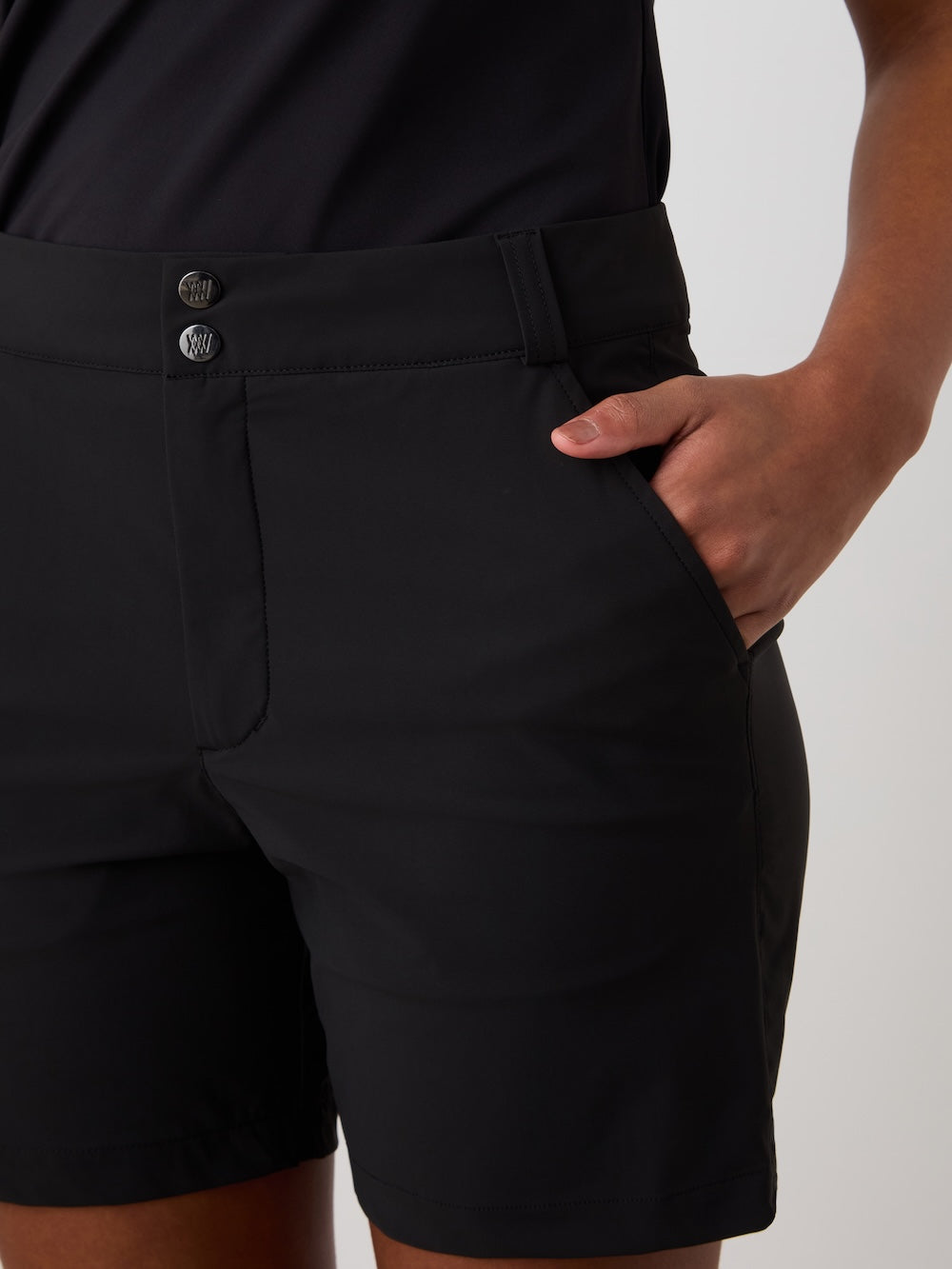 A woman wears We Norwegians Albatross Shorts, featuring black water-repellent fabric, front buttons, a waistband, and 4-way stretch for comfort and style, standing with one hand in her pocket.