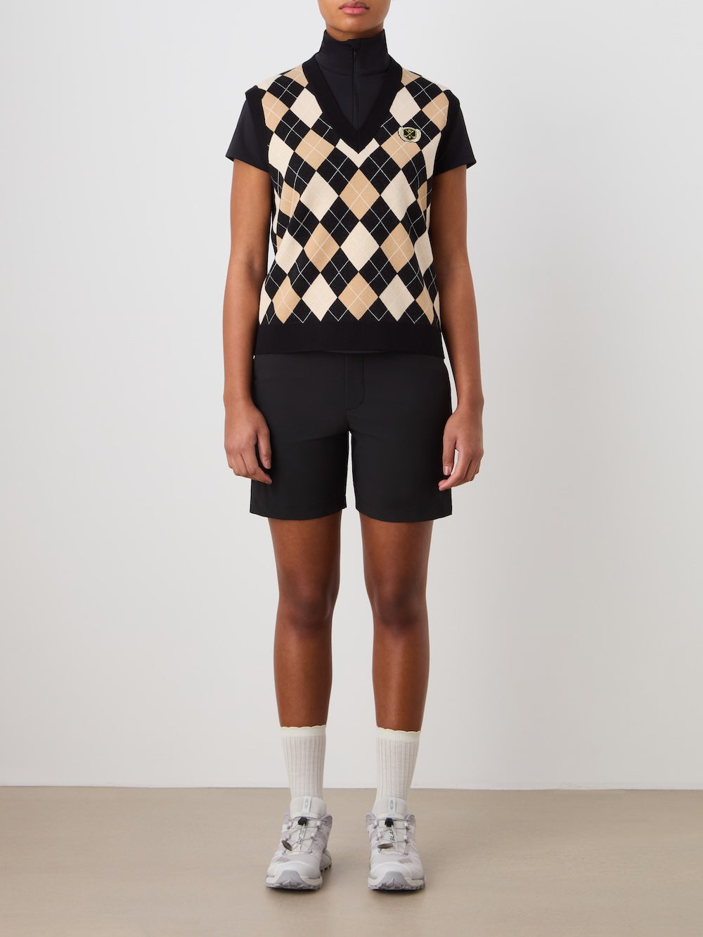 A person stands against a plain background wearing the We Norwegians Fairway Vest Women over a black short-sleeve shirt, paired with black shorts, white socks, and gray sneakers. Their arms are relaxed by their sides.