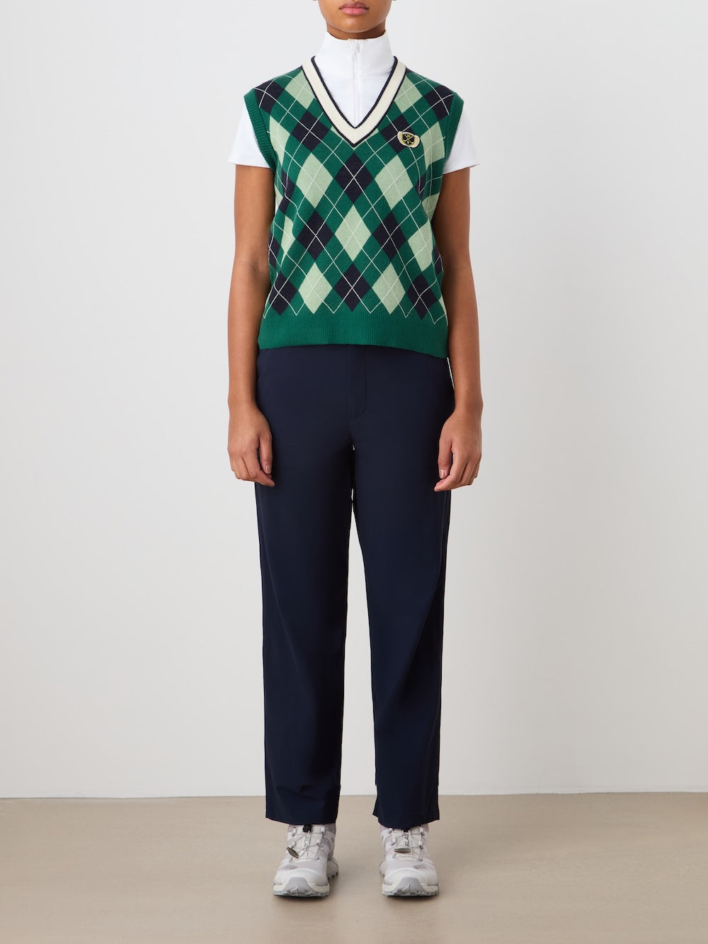 A person stands against a plain background wearing the We Norwegians Fairway Vest Women in green over a white turtleneck, paired with navy trousers and white sneakers. Their arms are relaxed at their sides.