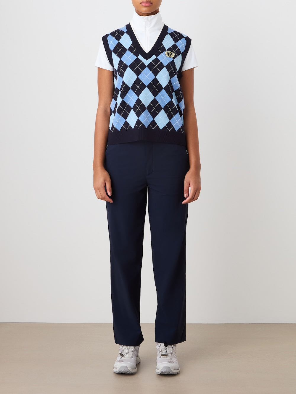 A person stands against a plain background, wearing the We Norwegians Fairway Vest Women in navy and light blue argyle over a white turtleneck, paired with navy pants and gray sneakers. Their arms rest relaxed at their sides.