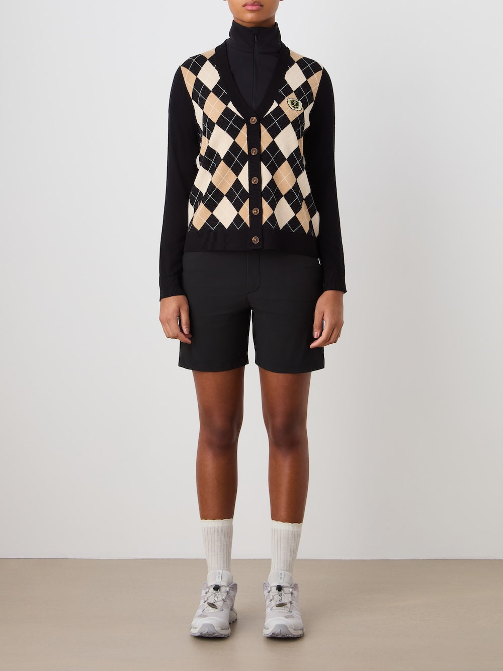 A person stands against a plain background wearing the We Norwegians Fairway Cardigan Women, paired with black shorts, white crew socks, and gray sneakers. Their arms rest at their sides.