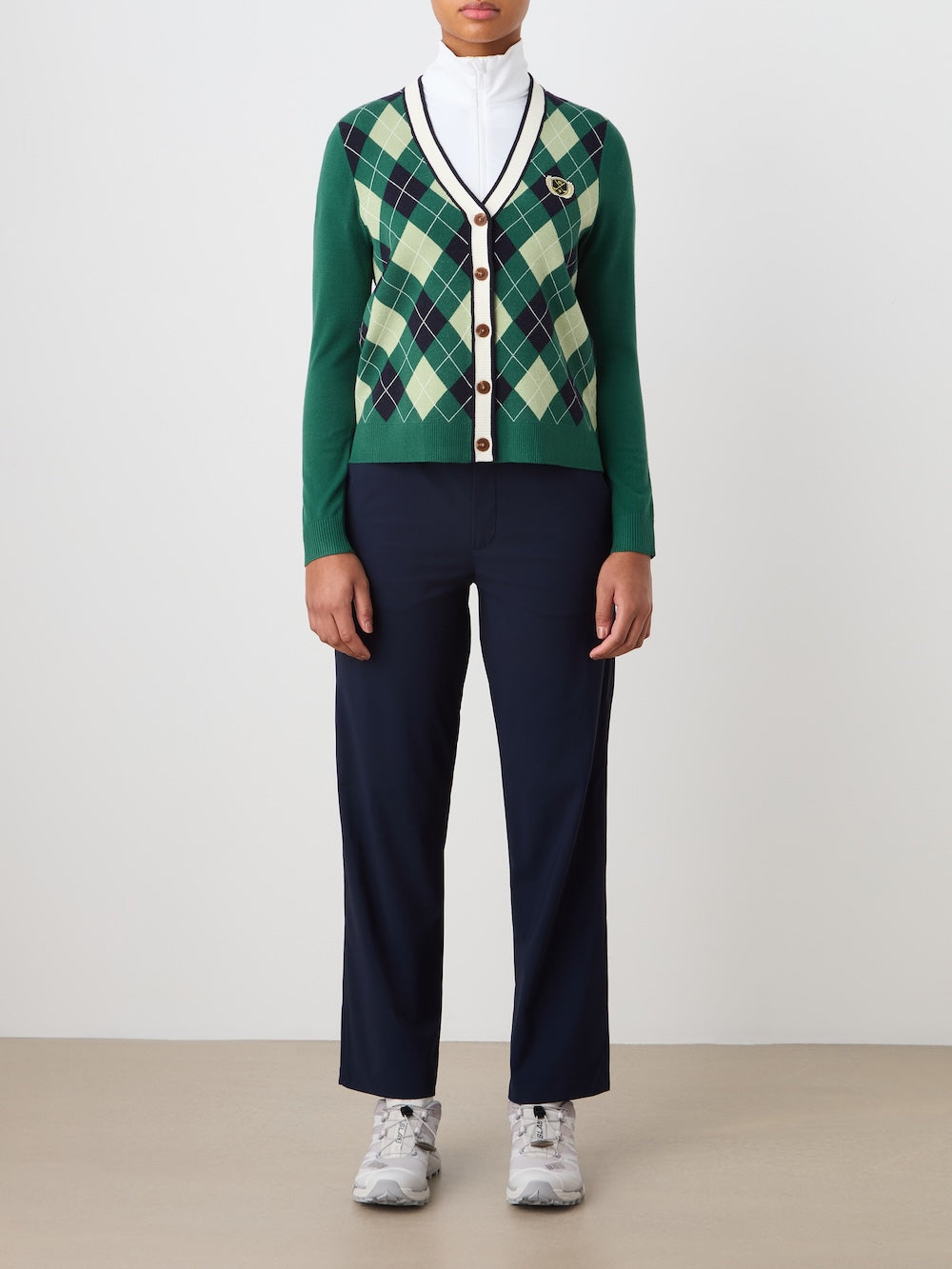 A person stands against a plain background wearing a We Norwegians Fairway Cardigan Women in green, navy, and cream over a white turtleneck, navy pants, and light gray sneakers. Their hands are by their sides.