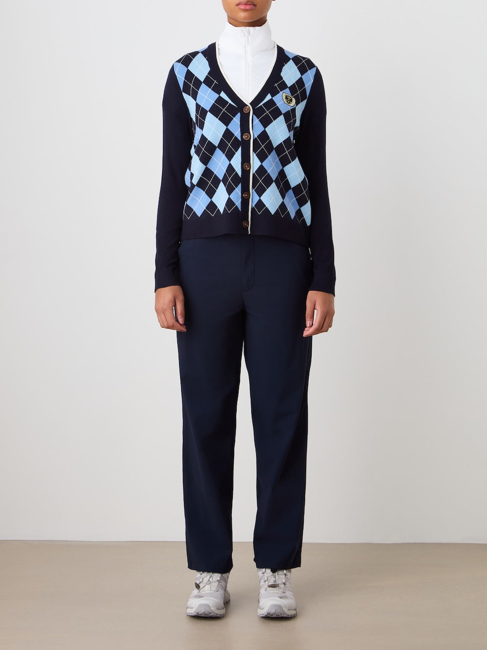 A person stands against a plain background wearing the We Norwegians Fairway Cardigan Women, featuring light blue and white argyle over a white turtleneck, dark navy pants, and white sneakers. Their hands are by their sides with their face partially visible.