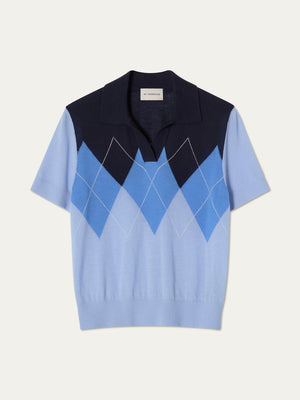 The Bandit Polo Women by We Norwegians is a women's oversized 100% cashmere short-sleeve knit polo, featuring a collared design and striking blue-and-white argyle front against a plain backdrop.