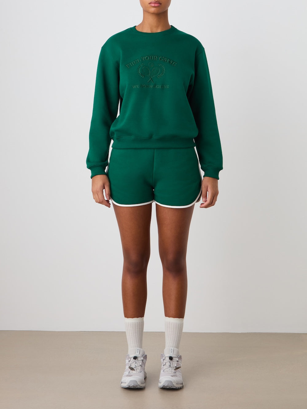 A person, face not shown, stands against a plain background wearing the We Norwegians Bounce Sweater Women and matching dark green shorts with tennis embroidery, paired with white socks and sneakers for a casual, sporty look.