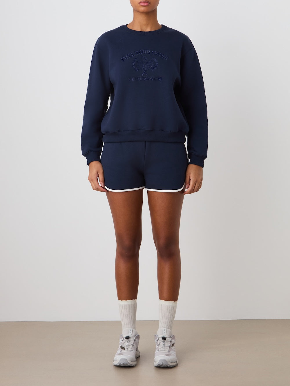 A person stands against a plain background wearing the We Norwegians Bounce Sweater Women in navy. They pair it with matching navy shorts with white trim, white socks, and gray sneakers. Their arms are relaxed and their face is not visible.