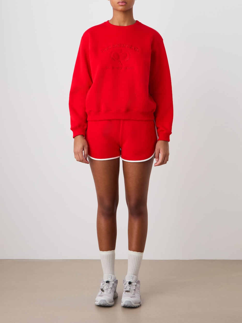 A person stands against a plain background wearing the We Norwegians Bounce Sweater Women in red, paired with matching red shorts with white trim, white crew socks, and gray athletic shoes. Their arms are relaxed at their sides.