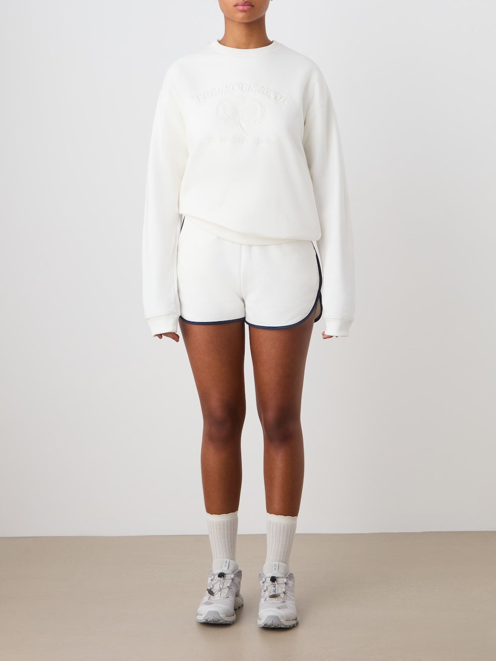 A person stands against a plain background wearing the We Norwegians Bounce Sweater Women, paired with white athletic shorts with dark trim, white socks, and sneakers. Arms are relaxed at their sides; subtle tennis embroidery accents the outfit.