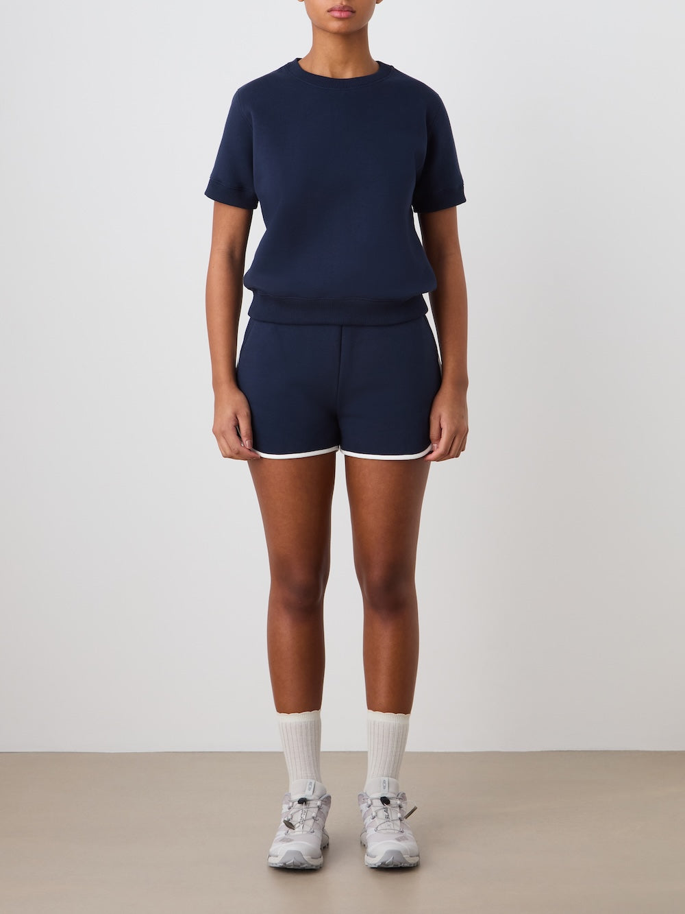 A person wears the We Norwegians Bounce Top Women, paired with matching navy shorts with white trim, white socks, and gray sneakers. They stand against a plain background, arms at their sides, face partially out of frame.