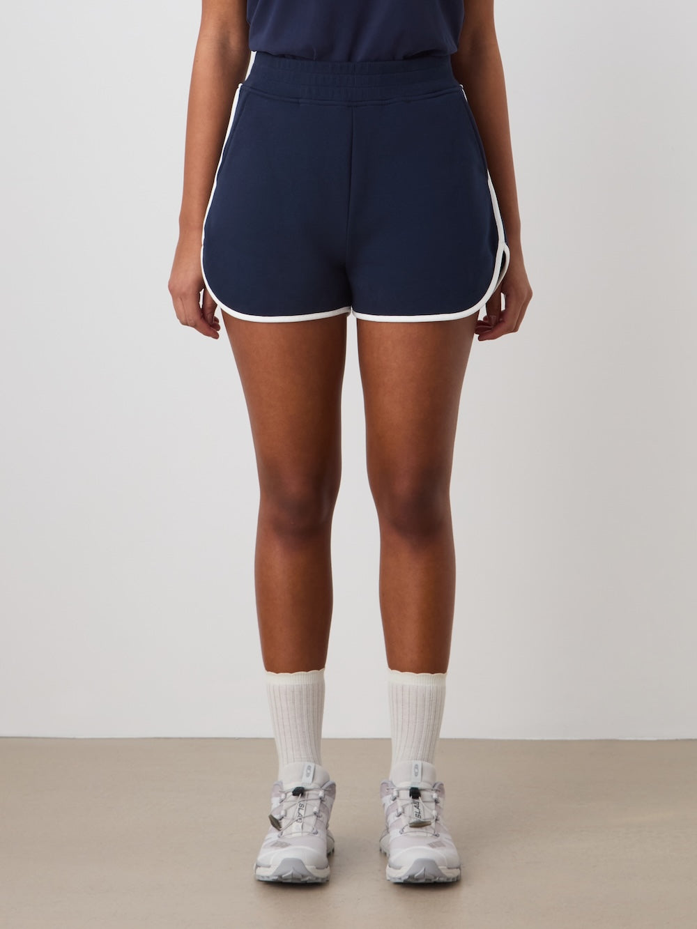 A person stands against a white wall wearing We Norwegians Bounce Shorts Women in navy with white trim, paired with white crew socks and gray sneakers. The 70s tennis-inspired outfit is shown from the neck down, arms at their sides.