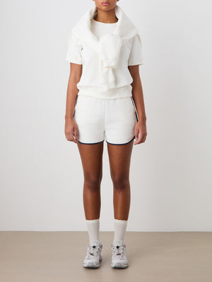 A person stands against a plain background wearing the We Norwegians Bounce Top Women, paired with white shorts featuring dark trim, a cotton sweatshirt draped over their shoulders, white socks, and athletic shoes. Their face is partially out of frame.