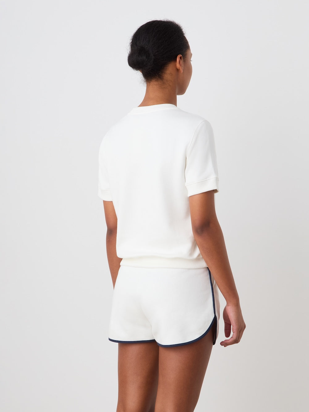 A woman with dark hair in a bun faces away, wearing a white short-sleeve top and We Norwegians Bounce Shorts Women—retro tennis shorts with dark trim—evoking classic 70s style against a plain light background.