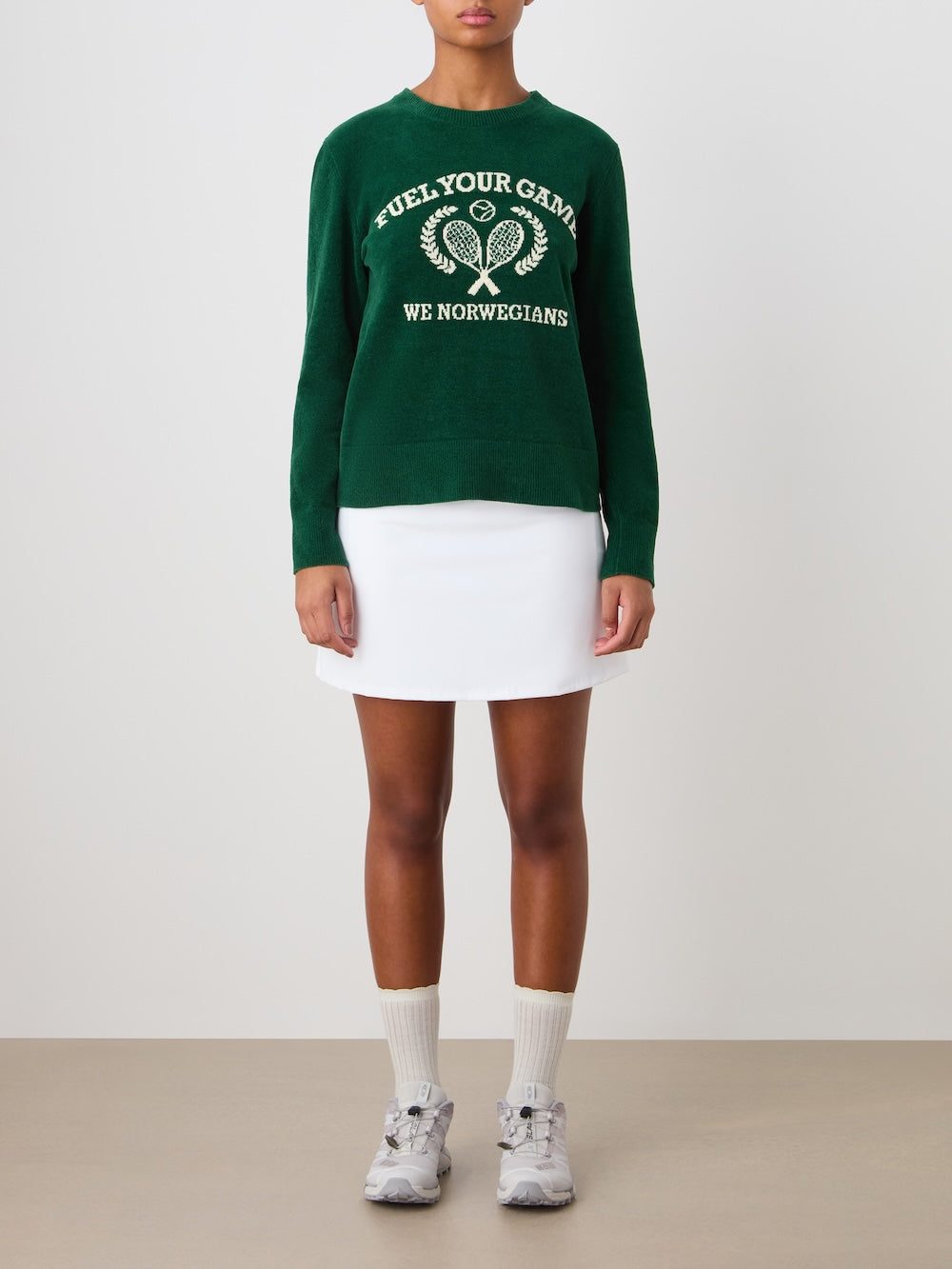 A person stands against a plain background wearing the We Norwegians Deuce Chenille Sweater for women in green with an intarsia tennis motif and “Fuel Your Game” text, paired with a white skirt, socks, and gray sneakers. Made in Italy.