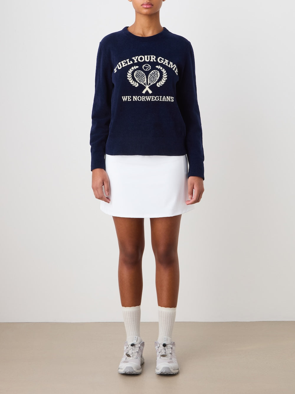 A person stands against a plain background wearing the We Norwegians Deuce Chenille Sweater Women, featuring a navy cotton chenille knit with an intarsia tennis motif and text, paired with a white skirt, socks, and sneakers. Hands rest by their sides.