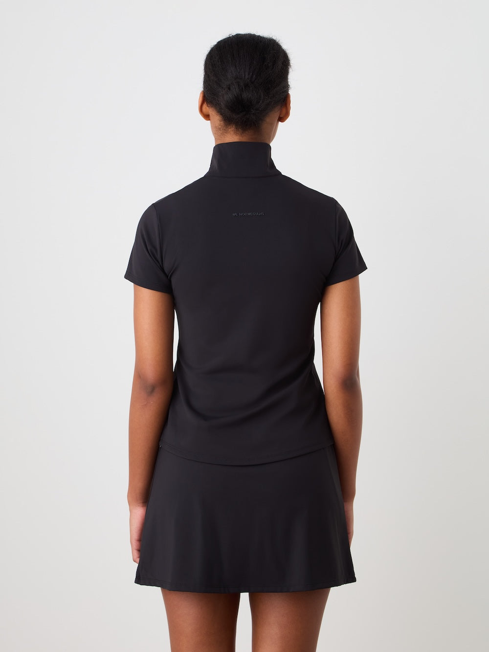 A woman faces away from the camera in a We Norwegians Honor Zipped Top Women—a short-sleeved black performance training top with a high collar—paired with a black skirt, set against a plain white background.