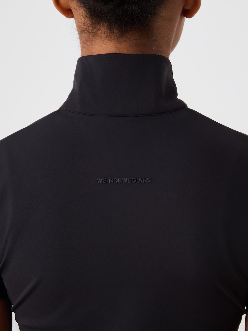 Close-up of a woman’s upper back and neck wearing the Honor Zipped Top by We Norwegians, a black high-collared performance shirt with “WE NORWEGIANS” subtly printed below the collar on the back.
