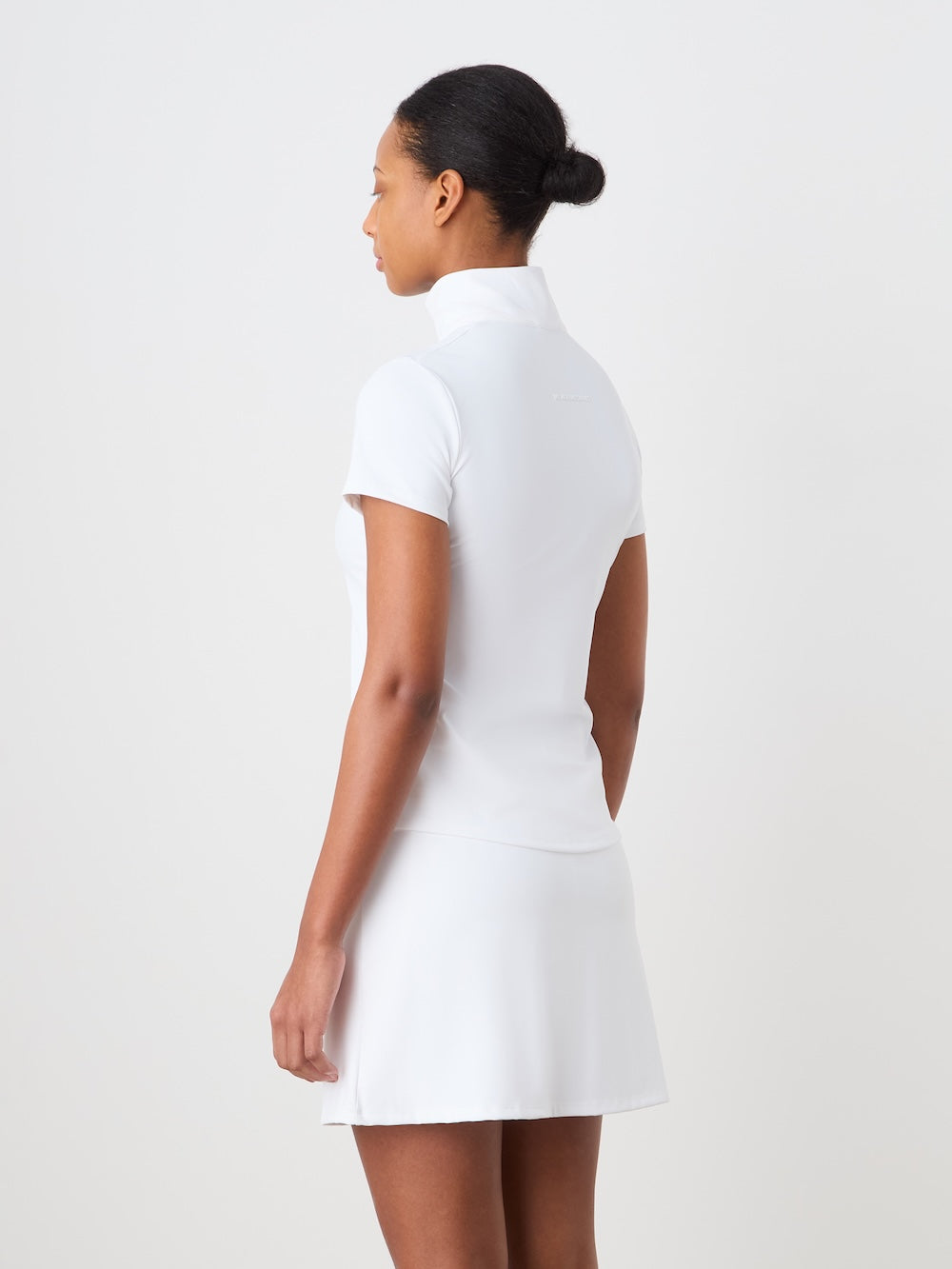 A woman faces away, wearing the We Norwegians Honor Zipped Top for women, styled with a matching skirt. Her hair is in a low bun, set against a plain light-colored background.