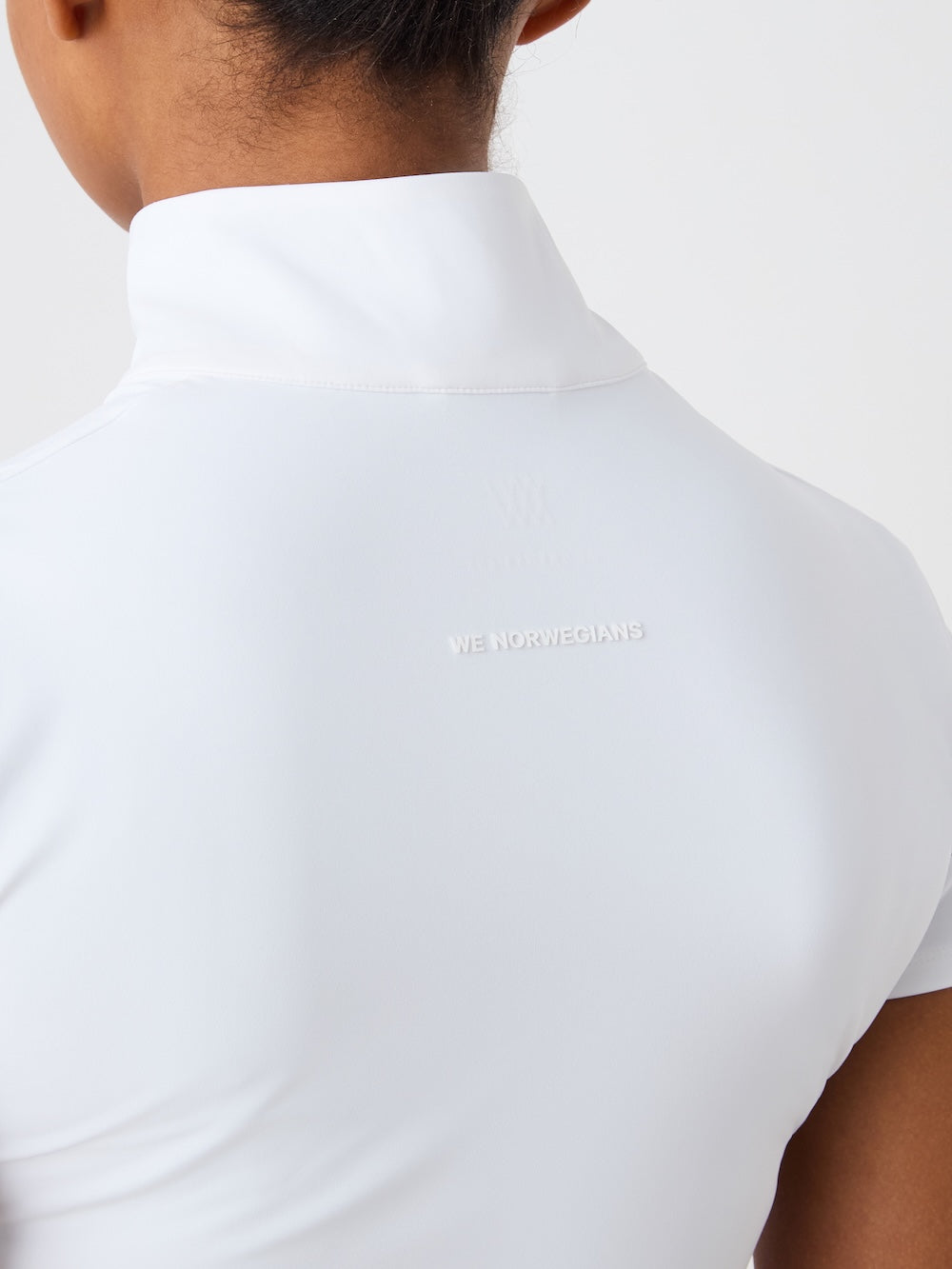 The image shows the back of a person wearing the We Norwegians Honor Zipped Top Women in white, with "WE NORWEGIANS" subtly printed below the collar. The background is plain and light-colored.