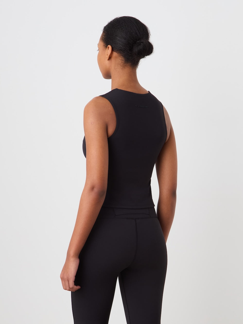 A woman with her hair in a bun stands facing away, wearing the fitted black We Norwegians Honor Top Women and black leggings against a plain white background.