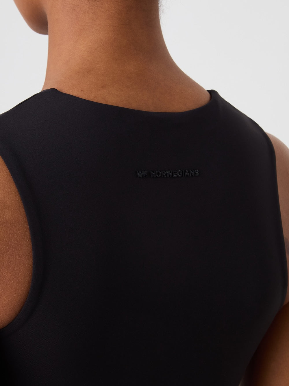 Close-up of the back of a person wearing the We Norwegians Honor Top Women, a cropped sleeveless style with subtle "WE NORWEGIANS" text near the neckline. The high-performance fabric contrasts against the plain, light background.