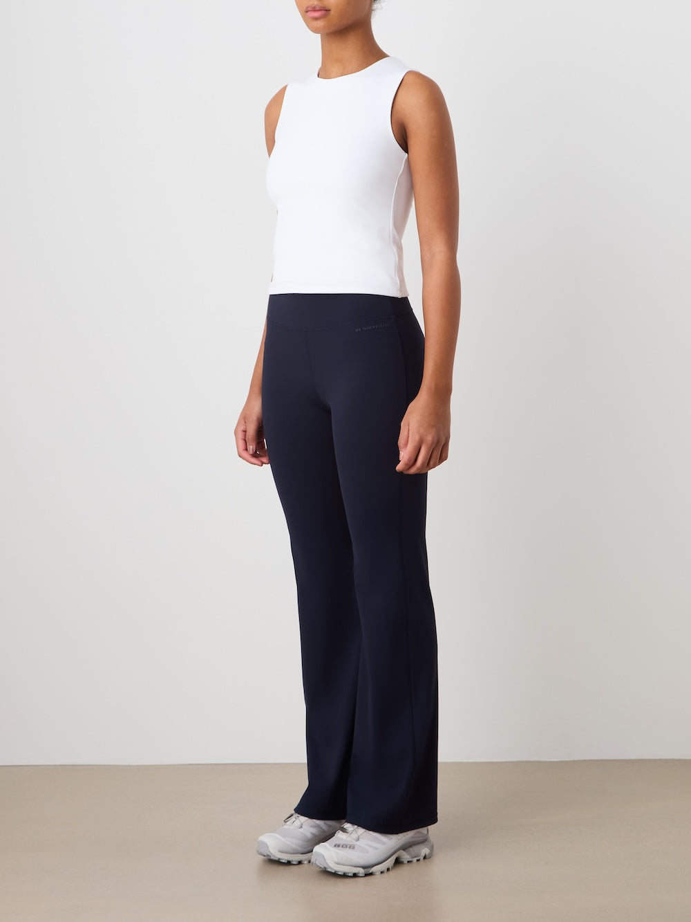 A person wearing the We Norwegians Honor Top Women, paired with navy blue flared pants and white sneakers, stands head to toe against a plain off-white wall on a beige floor, facing slightly left.