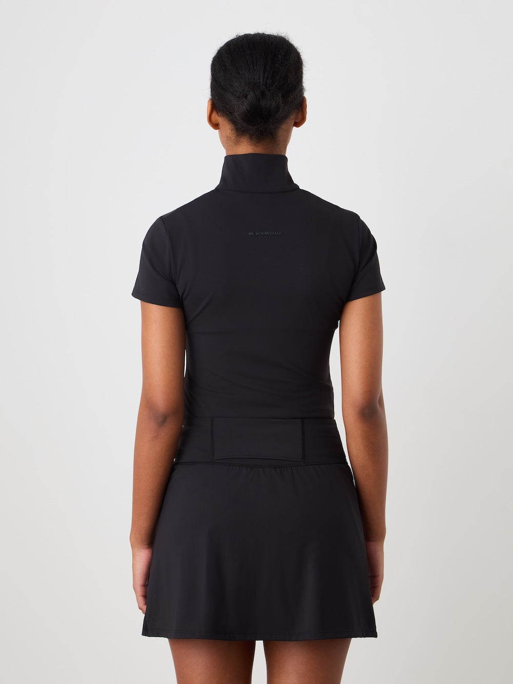 A person faces away from the camera, wearing the We Norwegians Honor Skirt Women and a short-sleeved black top with a high collar. Their hair is neatly tied back, and the background is plain white.