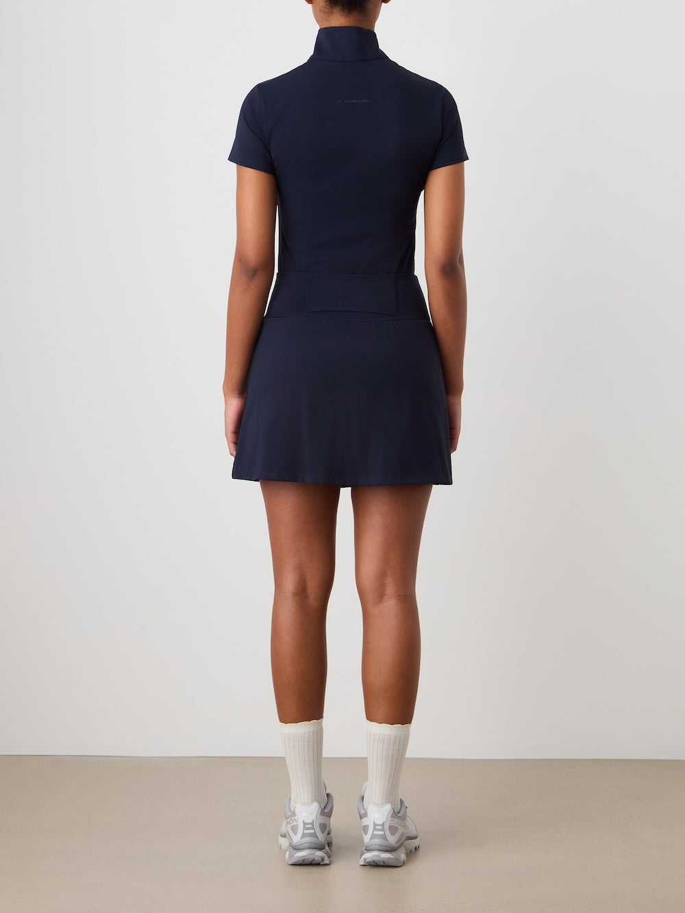 A woman faces away from the camera, wearing the We Norwegians Honor Skirt Women with built-in shorts, paired with white crew socks and sneakers, set against a plain light background.