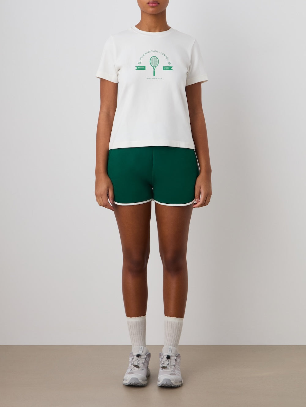 A person stands against a plain background wearing the We Norwegians Love Tee Women, green athletic shorts with white trim, white crew socks, and gray sneakers. Their arms are relaxed at their sides.