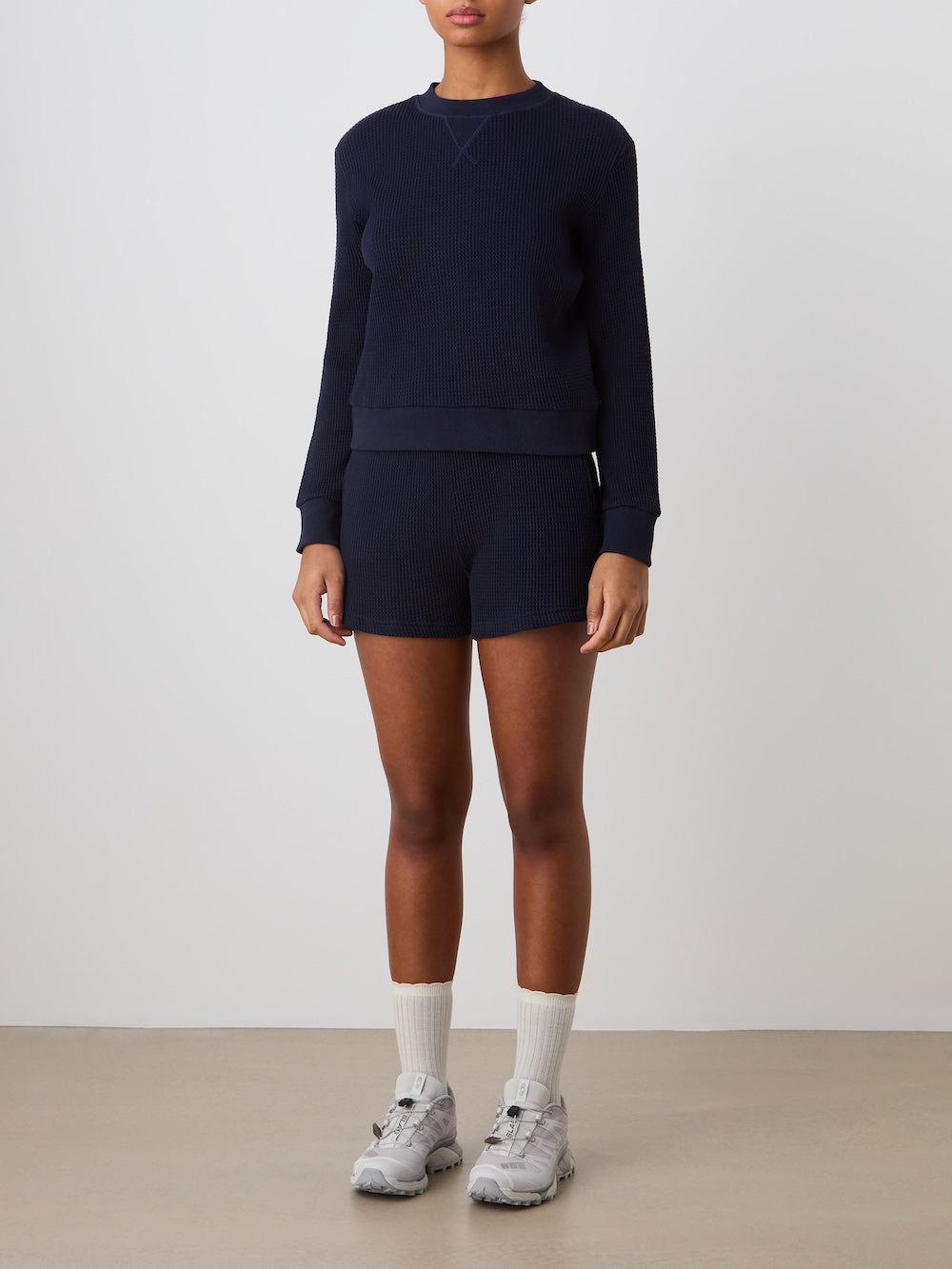 A person wearing the We Norwegians Waffle Crewneck Women in dark navy blue with matching shorts, white crew socks, and gray sneakers stands by a light-colored wall. Their face is not fully visible.